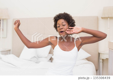 African American woman yawning and stretching on bed in bedroom with white linens and lamps African American woman yawning and stretching on bed in bedroom with white linens and lamps 135306439