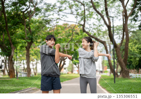Smiling asian man and woman in sportswear stretching arm together while standing on pavement in park Smiling asian man and woman in sportswear stretching arm together while standing on pavement in park 135306551
