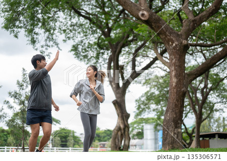 Asian man raising a hand and woman in sportswear smiling during a run or jogging on pavement in park 135306571
