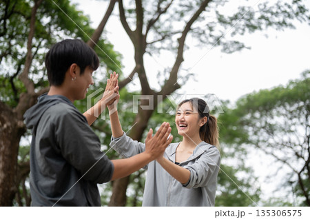 Asian man and woman in sportswear smiling as clapping hand after running or jogging together in park 135306575