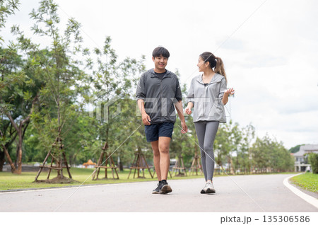 Asian woman in sportswear looking and talking with a man while walking together on pavement in park. 135306586