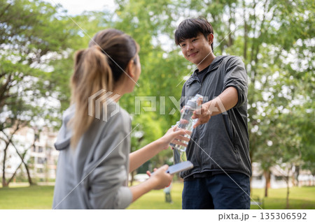 Asian man in sportswear holding a water bottle getting it or giving to a woman at park's courtyard. 135306592