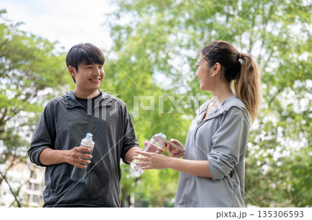 Asian man in sportswear holding water bottles and giving to a woman with phone at park's courtyard. Asian man in sportswear holding water bottles and giving to a woman with phone at park's courtyard. 135306593