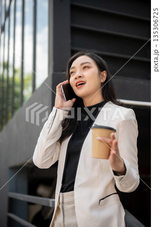 Smiling asian office worker businesswoman holding coffee talking on phone standing outside building 135306725