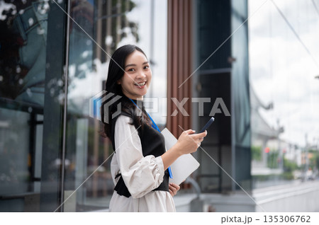 Smiling office worker or event staff holding folder looking away from phone standing out of building Smiling office worker or event staff holding folder looking away from phone standing out of building 135306762