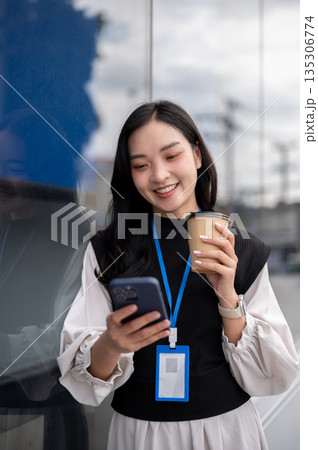 Female office worker or event staff holding coffee looking at phone while standing outside building 135306774