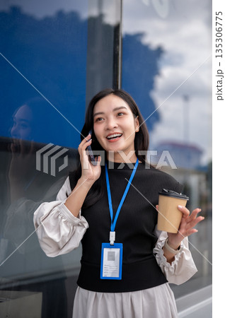Female office worker or event staff holding coffee talking on phone while standing outside building. 135306775