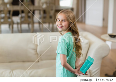 Female child standing behind cream sofa holding turquoise gift box in living room, copy space 135307032