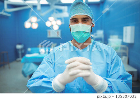 Male surgeon standing wearing blue scrubs and mask in operating room with surgical lights Male surgeon standing wearing blue scrubs and mask in operating room with surgical lights 135307035
