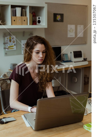 Twenty-something woman sitting and typing on laptop in home office with papers printer and fan 135307067