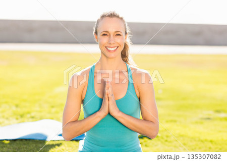 Woman in her 30s kneeling on grassy field beside blue yoga mat, wearing turquoise sportswear Woman in her 30s kneeling on grassy field beside blue yoga mat, wearing turquoise sportswear 135307082