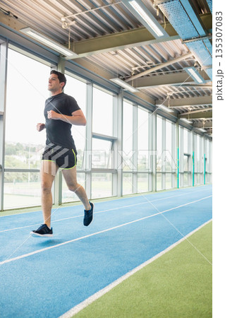 Male runner jogging along blue track inside training center by glass windows, copy space Male runner jogging along blue track inside training center by glass windows, copy space 135307083