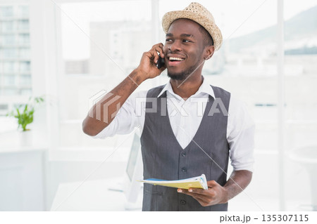 African American male in straw hat speaking on smartphone holding folder in office by PC monitor 135307115