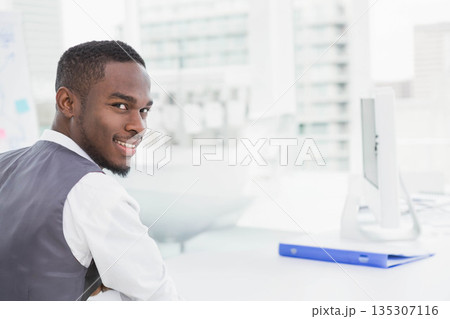 African American man sitting at white desk in office wearing vest viewing monitor and blue binder 135307116