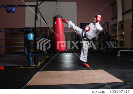 Male karate athlete wearing white uniform delivering side kick to punching bag in dojo, copy space Male karate athlete wearing white uniform delivering side kick to punching bag in dojo, copy space 135307117