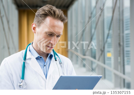 Male doctor wearing lab coat and stethoscope reading tablet in glass hospital corridor, copy space 135307155
