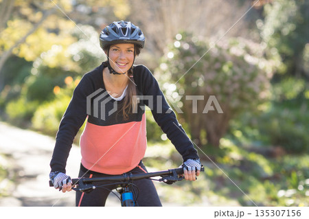 Adult woman in sportswear riding mountain bike along sunlit park trail with helmet and gloves 135307156
