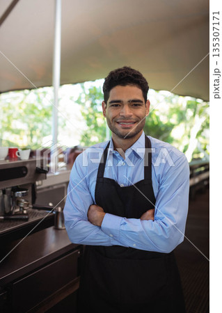 Man wearing black apron standing behind coffee counter at cafe with espresso machine, cups 135307171