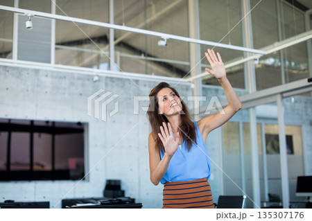 Woman gesturing toward virtual interface on glass partition in open-plan office with wall monitor 135307176