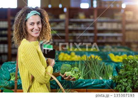 Woman wearing yellow sweater holding smartphone and basket selecting broccoli from crates at store 135307193