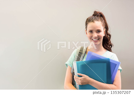 Teenage student standing against pale wall holding blue and teal binders, grey backpack, copy space 135307207