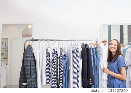 Woman organizing striped tops and denim on metal rack in retail boutique by window, copy space 135307209