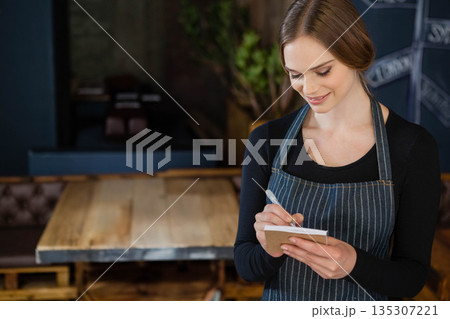 Female server standing beside table at restaurant writing order on notepad with pen, copy space Female server standing beside table at restaurant writing order on notepad with pen, copy space 135307221