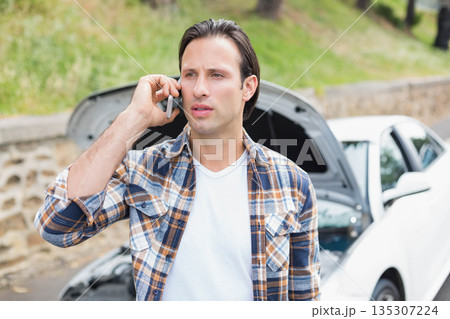 Man holding smartphone calling for help on roadside beside open-hood white sedan by low stone wall 135307224