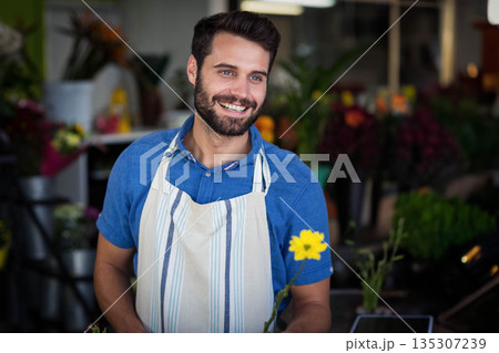 Male florist wearing striped apron behind flower shop counter holding yellow bloom and using tablet Male florist wearing striped apron behind flower shop counter holding yellow bloom and using tablet 135307239