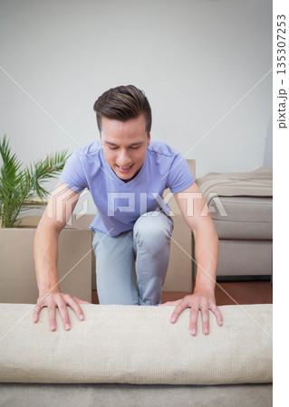 Man kneeling on floor in living room gripping beige sofa cushion with moving boxes and plant Man kneeling on floor in living room gripping beige sofa cushion with moving boxes and plant 135307253