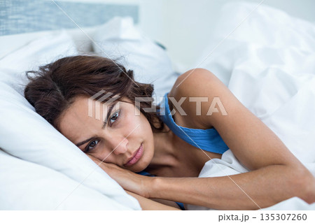 Mid adult woman lying on bed, wearing blue sleeveless top and resting head on white pillow Mid adult woman lying on bed, wearing blue sleeveless top and resting head on white pillow 135307260
