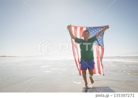 Boy running through shallow ocean water on beach holding American flag billowing under clear sky Boy running through shallow ocean water on beach holding American flag billowing under clear sky 135307271