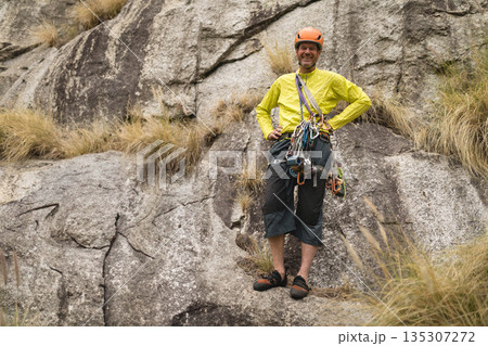 Senior male climber standing on rock cliff wearing yellow jacket and helmet with harness and rope 135307272
