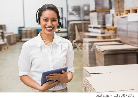 Woman wearing headset and shirt standing in warehouse holding tablet monitoring forklift and boxes Woman wearing headset and shirt standing in warehouse holding tablet monitoring forklift and boxes 135307273