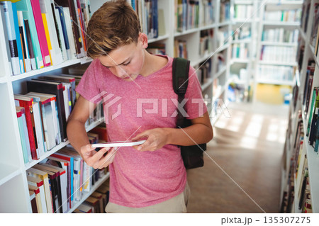 Teenage boy reading paperback book by white bookshelves in aisle with dark backpack, copy space 135307275
