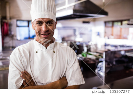 Mature chef standing in restaurant kitchen wearing uniform and toque with stainless steel counters 135307277