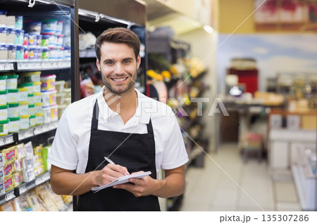 Male grocery employee wearing black apron over white shirt holding pen and notepad in grocery aisle Male grocery employee wearing black apron over white shirt holding pen and notepad in grocery aisle 135307286