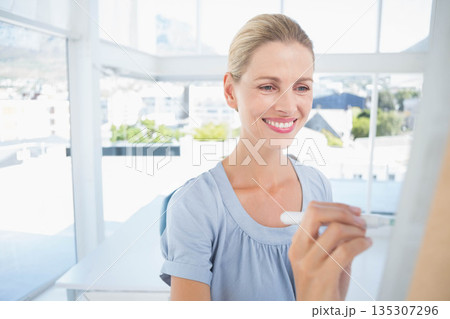 Middle-aged woman writing on whiteboard with marker at office desk with chair overlooking skyline Middle-aged woman writing on whiteboard with marker at office desk with chair overlooking skyline 135307296