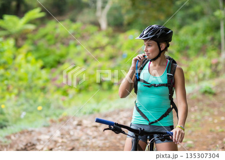 Female cyclist pausing on forest trail and sipping water from tube beside mountain bike, copy space Female cyclist pausing on forest trail and sipping water from tube beside mountain bike, copy space 135307304