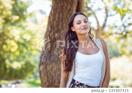 Woman leaning against tree trunk under sunlit foliage at park wearing floral skirt, copy space Woman leaning against tree trunk under sunlit foliage at park wearing floral skirt, copy space 135307315