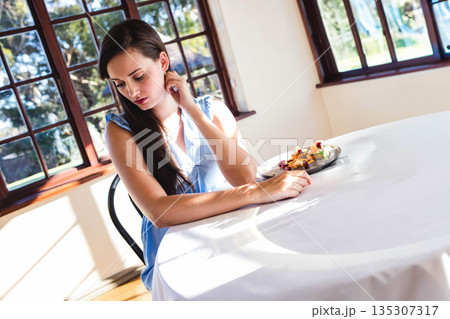 Woman sitting at round white cloth table by wooden windows, sampling pastries and fruit, copy space Woman sitting at round white cloth table by wooden windows, sampling pastries and fruit, copy space 135307317