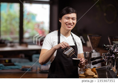 Asian woman barista in apron pouring milk with pitcher into cup in café with espresso machine 135307351