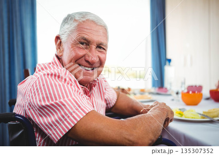 Senior man wearing pink polo shirt sitting in wheelchair at dining table eating eggs toast bowls Senior man wearing pink polo shirt sitting in wheelchair at dining table eating eggs toast bowls 135307352