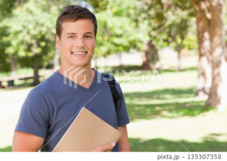 Male student wearing blue T-shirt on campus lawn, holding folder and backpack under sunlight 135307358