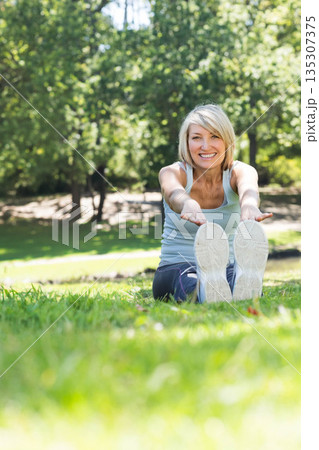 Middle-aged woman sitting on grass by pond in park touching toes while wearing white athletic shoes Middle-aged woman sitting on grass by pond in park touching toes while wearing white athletic shoes 135307375