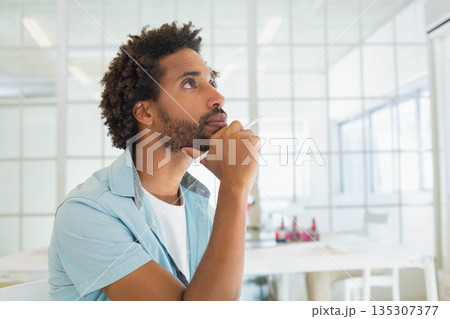 Mid adult African American man holding stylus pen, gazing upward at desk in office, copy space 135307377