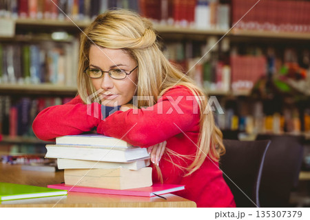 Female student leaning on textbooks at library table while wearing red sweater and eyeglasses 135307379