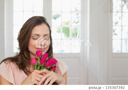 Mid adult woman standing at doorway holding pink roses close to face, copy space 135307392