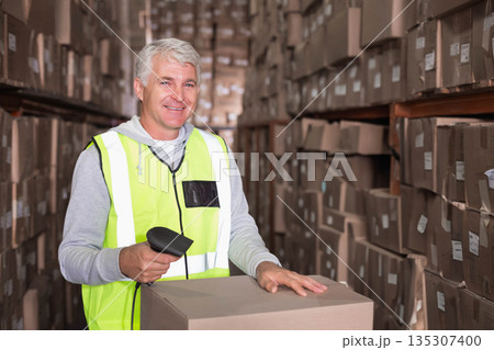 Male warehouse worker scanning box with barcode scanner in narrow aisle wearing neon safety vest 135307400