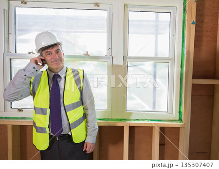 Senior man inspecting window frame inside building with hard hat safety vest and smartphone 135307407